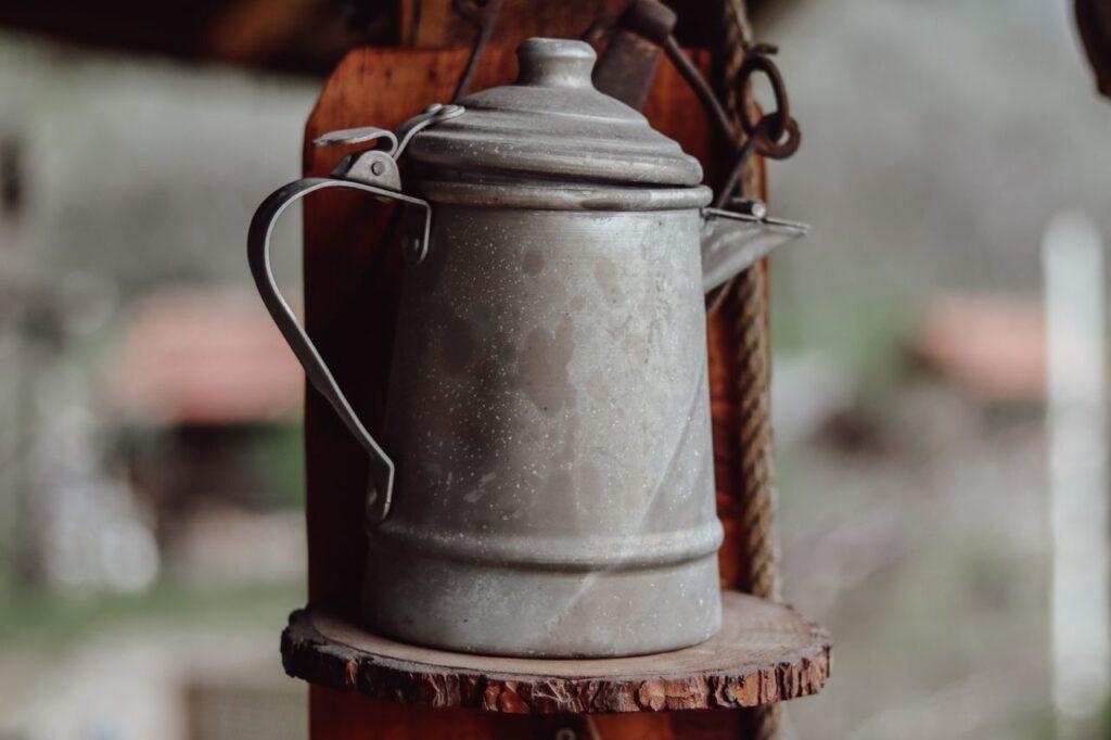 Rustic metal kettle with handle and spout displayed on a wooden shelf.
